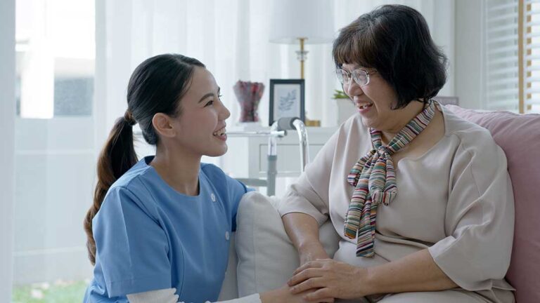 An older adult uses a tablet at a table while a caregiver points to the screen.